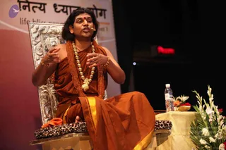 A man seated on an ornate silver throne-style chair speaks during a talk on a stage. He wears saffron-orange robes with a gold-bordered cloth, a long rudraksha mala and a heavy gold ceremonial necklace, plus gold bangles and a red bindi. He gestures with both hands while a small clip-on microphone is attached to his collar. A leopard-print cushion rests on the chair, and a side table holds a water bottle, a small offering bowl and white and red flowers. A banner behind him reads Nithya Dhyana Yoga. a person is seated centrally, delivering a discourse to an unseen audience.