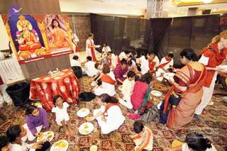 A photograph of a seated communal meal in a hall with a patterned carpet. A large printed banner on the left shows a goddess seated in red and gold with a golden crown and a man in orange robes with a rudraksha mala, labelled Devi Annapoorani and Paramahamsa Nithyananda. Many people sit on the floor eating from white plates, including children in white and orange. Volunteers in white with orange shawls serve food. A red-draped table holds a small offering tray. Dark drapes line the back wall.