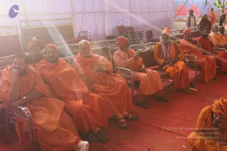 A photograph of a seated group of men in saffron-orange robes inside a tent with white draped walls and a red carpet. Several sit on metal-framed chairs with maroon cushions, some wearing orange or red turbans and caps, one with a white shawl. A man in the foreground left holds his chin in his hand, and a figure in the lower right corner sits on the floor in orange with a patterned head wrap. Behind them, a few standing figures wear orange and white, and sunlight streams in from the upper left. No identifiable a person candidate is clearly visible.