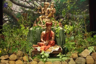 A seated man in saffron-orange robes and a red turban sits on a tiger-skin-draped wooden seat in a lush, staged forest setting of green plants, leafy branches and rocks. Long braided hair falls over his shoulders, and he wears a rudraksha-style necklace, armbands and bangles while smiling with hands folded in his lap. Behind him stands a large golden murti of a deity group, a seated figure with a raised hand and a standing consort, with smaller golden figures at its base and green garlands draped over a green cloth. A banyan-like tree canopy forms the backdrop.