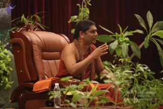 A man seated in a brown leather armchair with carved wooden armrests, speaking and gesturing with his right hand raised. He wears a rust-orange robe draped over his body with a tan-and-saffron cloth at his waist, and a long necklace of brown rudraksha beads hangs on his chest. A small lapel microphone is clipped near his mouth. Green leafy plants surround the chair, and an orange towel lies on the armrest beside a clear water bottle on a low table. Dark red drapes hang behind him. Swamiji (probable): long dark swept-back hair, saffron robe and rudraksha mala visible, seated as a teacher giving satsang.