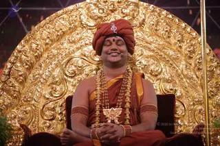 A seated man with closed eyes and a faint smile sits in front of an ornate gold repoussé backdrop with swirling floral carving. He wears a deep red turban with a small silver emblem, a red and gold robe, a rudraksha-style mala and layered gold ceremonial necklaces with a gold pendant, and gold bangles with red beads on both arms. A small red bindi is on his forehead and a clip microphone is at his face. Swamiji (probable) with long-established presentation cues: red-saffron turban, ornate gold necklaces and malas, and a throne-like seat, seated as a teacher in a discourse setting.