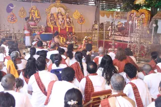 A photograph of a large ceremonial hall during a ritual. In the foreground, many seated devotees in white clothes with red-and-white striped shoulder cloths face the front, with a few heads shaved and some with long braids. At the front, a priest in a white dhoti stands at left beside a decorated altar with a deity murti draped in garlands, and brass oil lamps are lit along the stage. A central seated figure in a saffron garment sits on a raised red-draped platform beneath a golden canopy and floral arch, with musicians playing instruments and a sacred fire pit area near the front. Tall brass lamp stands and ornate gold hangings frame the scene.