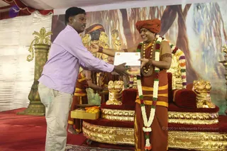 A man in a lavender striped shirt and cream trousers stands at left, extending a white certificate in a clear sleeve toward a seated figure at right. The seated person wears an orange-red turban, an orange-red robe with a yellow band, and multiple long garlands in white, red and green, plus beaded necklaces, and is seated on an ornate gold-and-red throne with carved lion-head armrests. Behind them a painted backdrop shows a large deity murti with a golden crown, and a fruit tray sits on the stage. Other attendants in orange are partly visible.