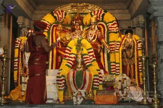 A temple shrine decorated with yellow and red-white marigold garlands shows gilded deity murtis of Shiva and Parvati on an ornate altar. In the foreground, a priest in a deep maroon dhoti and matching shawl with a red turban stands with his back to the camera, holding a lit lamp toward the deities. A kalasha stands before the altar. Brass oil lamps flank the sides. Beside the shrine, a statue of a sage in a tiger-skin garment holds a staff. No living person is visible beyond the priest, and no a person candidate is clearly present.