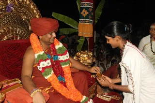 A seated man in orange robes and an orange turban sits on an ornate gold-and-red throne, smiling, wearing a large garland of orange marigolds with green and red flower sections and a blue bead strand. He reaches out to accept a small card or booklet from a young woman in a white sari with a red border, who wears a red-beaded necklace and a hair clip and bends toward him. Behind them a crowd of people in white and orange sits in the dark. A banana-leaf decoration and a red-and-gold patterned hanging appear in the background, with brass items and printed booklets on the lower left. Swamiji (probable) is visible: the seated man wears saffron/orange clothing, a turban and a garland. A man in white with a beaded necklace and forehead markings stands at the right edge.