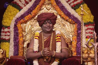 A seated man in a maroon-red turban and maroon-red robe sits on an ornate gold throne decorated with garlands of marigolds in orange, yellow, purple and white. He wears a rudraksha mala, a gold necklace, gold armbands and bangles, and a small bindi, and holds his palms together in front of his chest with eyes softly closed. A golden staff topped with a bird figure stands to the right. A small water bottle sits at the lower left. Watermarked archive photograph of an event.