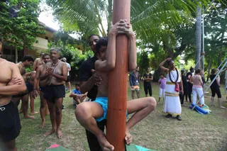 Outdoor photo on grass under palm trees at a Nithyanandoham session in Phuket. A boy in blue swim shorts clings to a tall wooden pole, legs wrapped around it and arms gripping it high. Behind him an adult in a black shirt steadies him with one hand. Nearby, shirtless men in black shorts stand with hands folded or arms crossed, and a woman in a white dress with gold border and a red bag stands with one arm raised. A yellow-and-white building and palm trees fill the background, with more participants in dark and pink clothing further back.