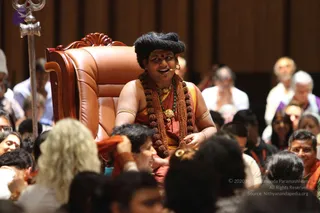 A seated figure in a large carved brown leather throne chair smiles and speaks into a headset microphone during a hall gathering. He wears a red-orange garment, multiple long rudraksha malas across his chest, a green bead necklace with a gold pendant, gold armlets and a red-orange tilak on the forehead, with dark hair swept up. A silver trident stands to the left. Audience members, many seated and some wearing orange or saffron, fill the foreground and rows behind him, and a woman with curly blonde hair is visible at left.