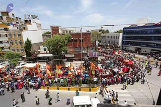 An elevated view of a busy city street during a procession. Dense crowds of people in colourful clothing fill the road and pavements, many in saffron, red and pink. Tall orange, yellow and red flags on poles rise above the crowd, and decorated carts or palanquin-like vehicles with red draped canopies move through the middle of the street. Multi-storey buildings, a red-brick structure, a small tree and parked white cars line the scene under a clear sky. Signboards on the buildings are visible, including one reading Emotions.