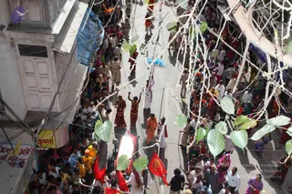 Elevated view of a dense street procession in an Indian town, with crowds packed along both sides of the road under a canopy of strung white flowers and large green leaves. Two men in saffron-orange clothing walk near the center, one with a small blue umbrella held over him. Uniformed police officers line the route. A white vehicle with red flags in front moves through the crowd. Many women in colourful saris watch from the sidewalks and balconies, and a shop sign with Hindi lettering is visible at left.