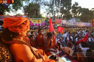 Outdoor festival stage at dusk with a crowd behind yellow barricades and red flags. In the foreground, a sadhu in an orange turban and gold-and-rudraksha necklaces sits from behind on a leopard-print seat, facing the stage. Beside him, several saffron-robed sadhus sit, one playing a drum (dhol-style) with his hands. A seated police officer in khaki uniform stands nearby. Banners and a red-and-pink banner reading KHAMA hang in the background, with trees and light poles above. Setting suggests a procession or ritual gathering with sadhus.