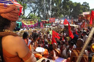 Outdoor festival procession crowd seen from behind a foreground figure in orange sannyasi attire with an orange turban and rudraksha beads, standing near the front of a barricade. A woman in orange with a long braid and beaded necklaces plays a small hand drum. Around them, saffron-robed sadhus, a bald monk in orange, and many devotees and onlookers, some in white, fill the road. Red flags on poles wave above the crowd, and a high-ranking officer in khaki uniform stands at the right. Behind, a canopied chariot-like cart, an orange and red decorated structure, colourful banners and tents line the road under trees. A yellow banner reads 'ARMA' with an animal motif, and a red placard carries a portrait. No identifiable a person figure is clearly visible.