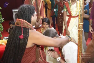 A photograph of a long-haired figure in a rust-coloured saffron-toned robe with a red and white beaded mala draped over the shoulder, reaching forward to lay a hand on the head of a kneeling person in white. The kneeling person has grey hair in a bun and wears white clothing with a gold wristwatch visible. Nearby, a woman in a cream sari with a red border and gold necklaces stands watching, and another woman in a cream and gold sari stands behind the kneeling figure. Other people in ethnic dress, including green and maroon garments, are visible in the background on a red carpeted floor beside a gold-framed pillar and green plants.