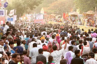 A dense crowd of many hundreds of people fills a wide city street during a religious procession, shot in warm late-afternoon light. Men in white shirts, kurtas and checked shirts walk among women in pink, purple and maroon saris and headscarves, and several men wear orange and saffron turbans. Behind them, decorated floats and palanquins with yellow and gold canopies and red and orange flags move along the road, and roadside barriers line the route. Hand-painted banners and signs stand along the street, and one man in the foreground raises his arm holding a phone or small object.