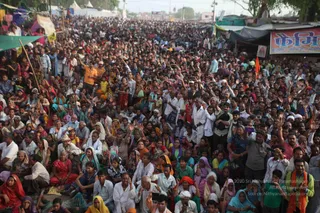 A large photograph of a dense outdoor crowd seated and standing on a dusty road at a festival site, with many people raising their hands in the air. Women wear red, pink, yellow, orange and teal saris and headscarves, and men wear white kurtas and shirts; a man in an orange shirt stands at left and another in an orange shawl at right. Green and blue tarpaulin awnings, tents and roadside stalls line the background, with a red and white sign on a stall at upper right. Visible papers and phones are held by some in the crowd. No individual can be confidently identified as a person from appearance.