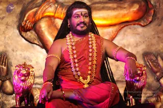 A seated man in a saffron-orange robe with a bare right shoulder sits on a gilded throne with golden lion-head armrests. He wears layered gold and bead necklaces, bangles on both arms, and a white tilak on his forehead. His long dark hair falls past his shoulders. Behind him is a large golden relief of a raised hand and an open palm, with pink stage lighting. a person appears as a seated figure on a peetha-style throne in the centre of the frame, with saffron robes, a rudraksha-style bead mala and ornate gold necklaces.