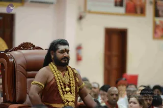 A bearded man with long dark hair and a white vertical tilak on his forehead sits on an ornate brown leather throne-style chair, wearing a rust-red shawl with multiple strands of heavy gold ceremonial necklaces, gold armlets and a gold waist belt. Swamiji (probable) is indicated by the long hair, ornate gold adornments and rudraksha-style ceremonial dress, and he is seated in the role of the presiding teacher. Behind him, a seated audience fills the hall, some in white with tilaks and others in plain clothes, with framed photographs on the wall and a wooden door in the background.