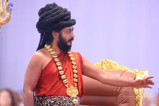 A man stands on a stage in front of a gold-and-brown ornate throne, extending his right arm toward the side. He wears a large dark dreadlocked crown-like hairstyle, a full beard, and an orange-red silk shawl draped over one shoulder, with a heavy gold bead necklace with a gold pendant and a leopard-print waist cloth. A gold trident is partly visible at the upper left, and blurred audience members sit in the foreground.