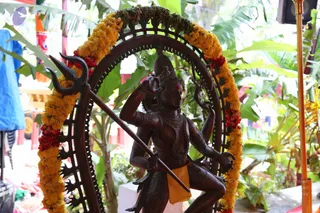A bronze-toned statue of Nataraja, the dancing Shiva, stands within a dark arched halo ringed with pointed flame motifs. The figure holds a trident in one hand and a drum-like object raised in another, wearing a bead necklace and an orange-yellow waist cloth. Yellow and red marigold garlands drape the arch. Behind it are green banana leaves and a plastic-wrapped stall or tent, and a brass pole stands at right.