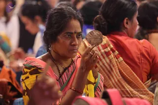 A woman in the foreground holds up a dried leaf between her fingers and looks at it intently. She wears a pink and yellow patterned sari with a striped green border, a red bangle, a small bindi, a nose stud, and a dark beaded mala around her neck. Behind her, other women in red and orange saris are seated among a crowd at what appears to be a gathering, with the dried leaf being the central object of attention.