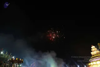 Night photograph of a fireworks burst in a dark sky above a festival ground. Red and white sparks fan out from the centre, with smoke drifting low across the lower left. On the right stands a tall, tiered decorative tower covered in illuminated flower garlands in white, yellow, pink and purple. A colourful striped canopy and string lights glow at the lower left, where a few small figures in the crowd are visible at the edge. The frame is dominated by the black night sky.