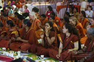 A large hall of seated women in rust-red and orange saffron saris and dresses with dark rudraksha malas around their necks, sitting on the floor in rows. Each has a banana leaf with food and small paper cups in front of them. Several women with gold trim on their clothes are eating or smiling, one in the centre lifts food to her mouth. Behind them, orange-draped tables and more seated devotees fill the background. The setting is a festive community meal gathering.