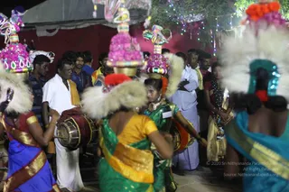 A night-time street celebration with a crowd of local men and women gathered around performers. Several women in the foreground wear saris in blue, green, yellow and teal, and carry tall decorated headpieces topped with pink flower arrangements, white feathered plumes and colourful painted figures. Drummers in white dhotis and a man beside them play large barrel drums, and a crowd in shirts and dhotis watches under strings of coloured fairy lights. A red tent and a horse-like painted figure appear at the top left. No individual is identifiable as a person from appearance.