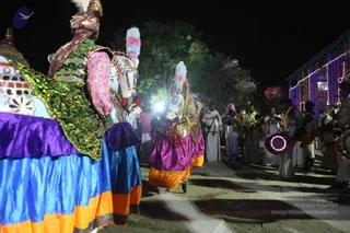 Night procession on a paved street lined with string lights on a building at right. Two dancers in elaborate costumes dominate the foreground: the left figure wears a sequined green and gold tunic over a blue and orange skirt with pink sleeves and a plumed headdress, while the centre dancer wears a purple and orange skirt, a sequined jacket and a white feathered headpiece with pink plumes, carrying a decorated drum. Behind them white-clad men in dhotis and white shirts play drums and wind instruments, and a drummer at right holds a lit pink-rimmed drum. A bright light flares near the centre. Spectators stand at the edge of the road under trees.