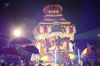 Night photograph of a festival procession. A large floral-covered ratha or chariot dominates the frame, its tiered superstructure wrapped in red, purple, white and yellow garlands with a golden kalasha-like crown on top. Several men stand on the platform in white dhotis and orange-saffron garments, wearing rudraksha malas and gold necklaces; the central figure has a tall crown-like headdress and ornate gold clothing, seated on a golden throne. Dark umbrellas with decorative fringe shade the platform, and a bright stage light glows at left. Dense crowd members are visible below.
