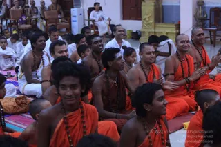 A photograph of a large seated assembly inside a temple hall during a satsang or puja gathering. In the foreground, several young men wear saffron-orange cloth draped over the shoulders and wooden rudraksha malas around their necks, smiling as they sit close together. Behind them, a few men in white ritual clothes and younger children in white and orange sit on mats and chairs. A golden shrine with a carved deity figure stands at the upper right, and a white air cooler and wooden chairs are visible at the back. The overall scene shows devotees gathered attentively in an ashram hall.