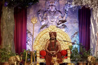 A photograph of a man seated cross-legged on an ornate golden throne with red cushions, in a stage setting for a discourse. He wears a rust-orange turban, rust-orange robes, a bare-chested upper body with gold armlets, and a long rudraksha mala with ceremonial gold pendants; his hands rest together in his lap. Behind him a large silver-toned relief of a multi-armed deity murti holding a book and other objects rises above purple drapery and potted green plants. A golden trident stands to his left, golden elephant figurines sit at the base, and a microphone stand is beside him. Swamiji (probable) based on the orange robes, turban and rudraksha mala with the page noting a discourse by Paramahamsa Nithyananda Swamiji.