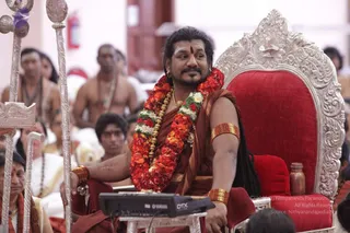A seated man sits on an ornate silver throne with a red velvet cushion, wearing a maroon-brown robe and a long multi-coloured garland of red and white flowers around his neck. He has gold bangles on his upper arm and wrists and a red forehead mark, and his long dark hair is swept back. Beside him stands a silver ceremonial staff. Behind him, several people sit in a hall with white walls, some bare-chested and others in white clothes, and a Yamaha DTX electronic drum unit sits on a small table in front. Swamiji (probable): the seated man has long swept-back hair, a multi-coloured flower garland, gold ornaments and a maroon robe on a throne, which are the qualifying cues.