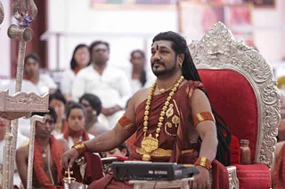A seated man in a silver ornate throne with a red cushion, wearing maroon-red draped cloth over the shoulder, heavy gold necklaces, gold earrings, red and gold armbands and a red tilak on his forehead. He holds a small silver ritual implement and sits beside a silver trident-topped staff and a Yamaha device on a table. Behind him, seated devotees in white and red-orange robes watch. The Swamiji (probable) cues are the long swept-back hair, gold ornaments and saffron-maroon ceremonial clothing, shown in an in-person satsang-style setting.