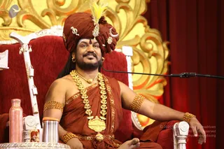 A man seated on a red velvet throne with carved white wooden armrests and a gold scrollwork backdrop. He wears a rust-brown turban with a yellow feather plume, a crescent ornament and jewelled pins, a brown draped garment, a heavy gold necklace with a large pendant, and beaded bracelets. He has a tilak on his forehead and sits before a microphone stand. A copper water bottle and a silver tumbler sit on a table beside him, with a white staff hung with bells leaning against the chair.