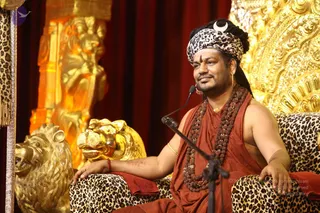 A man seated on an ornate leopard-print throne with gilded lion-head armrests and a golden carved backrest behind him. He wears a leopard-print turban with a crescent moon ornament, a rust-orange robe, multiple rudraksha mala necklaces, a gold chain, gold earrings, and a red-orange bead bracelet. A microphone on a stand is in front of him, indicating he is speaking, and a gold-toned carved column and dark red drapery frame the stage. Swamiji (probable): long swept-back hair, crescent-moon turban, rudraksha malas and ceremonial gold ornaments identify him as the presiding figure of the discourse setting.