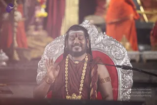 A photograph of a man seated on an ornate silver throne with a red velvet cushion, eyes closed, smiling, one hand raised in a blessing gesture. He wears a maroon-red wrap over his shoulders, a long multi-strand gold necklace, a gold chest pendant, gold armbands and bangles, and a white horizontal forehead mark with a red dot. A microphone on a stand is at his side. Blurred orange-robed attendants and a gold-and-red temple setting with a golden kalasha-style vessel stand behind him. The seated man has distinctive long swept-back hair, ornate gold necklaces and a crown-like setting of ceremonial dress, consistent with the probable identification of Swamiji seated for a discourse.