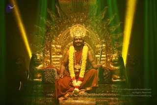 A seated man in orange-saffron robes sits on an ornate gilded throne with carved lion heads and a leopard-print cushion. He wears a crown-like golden headpiece, a long yellow and red garland, rudraksha beads and a gold ceremonial pendant, and holds a golden staff in his right hand. Green and yellow stage lights frame the scene behind the throne.