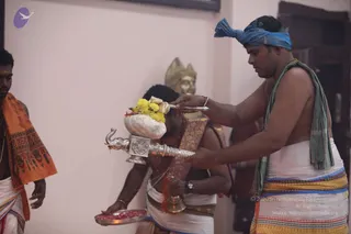 A temple ritual in a plain room with pale pink walls. A bare-chested priest in a blue headcloth and white dhoti with a striped border pours water from a silver elephant-spouted kalasha, its top decorated with yellow marigolds, over the head of a kneeling man in a white dhoti and red garland. Another attendant in an orange-and-saffron shawl stands at left holding a tray of red petals, and a gilt deity murti is visible behind. The scene shows an abhishekam-style blessing.