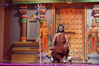 A seated man in a rust-red dhoti-style garment and matching shawl sits cross-legged on a tiger-striped cloth draped over a low wooden seat on a stage. He wears a long rudraksha mala with a large necklace, bangles, earrings and a tilak on his forehead, with long dark hair swept back. Beside him stands a tall silver trident with a small hand drum hanging from it and a leopard-print tassel. Behind him a wooden door with a lattice pattern is flanked by painted deity figures in orange and gold with green sashes, as in a temple set. The appearance of this seated man with saffron-red clothing, rudraksha mala and long hair is a person match, shown as a stage presentation with the trident and tiger-skin seat. He is the only visible person.