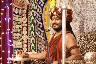A smiling man with a beard sits on a throne draped in leopard-print cloth, in front of an ornate silver backdrop and richly embroidered gold and red textile panels. He wears a rust-orange turban with gold ornaments, an orange-red robe, multiple rudraksha malas, and a gold necklace, with rudraksha bracelets on his wrists. A microphone stands before him and a small framed photograph sits on a side table at left, beside string lights. The figure's appearance matches the page's a person reference, but cues are only partially distinctive, so identity is uncertain.