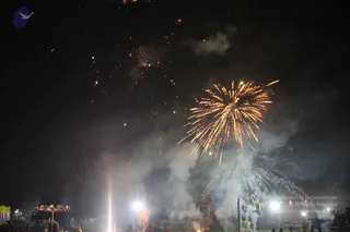 Night photograph of a fireworks display bursting over a dark sky, with a large golden-orange chrysanthemum burst at right and sparks falling downward. Smoke drifts through bright floodlights at ground level. Faint silhouettes of a few distant people stand near a lit structure and a tall tower or scaffold at the lower right, with a colourful illuminated stall or canopy at lower left. No individual figure can be identified clearly.