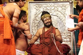 A photograph of a seated man in a rust-red robe with a yellow and black patterned headwrap, wearing rudraksha malas and a gold necklace, with eyes closed and smiling. He sits on a maroon cushioned seat before an ornate silver throne backrest. Around him, attendants in orange garments assist: one in orange with white horizontal body markings holds his hands, another shirtless devotee with white marks kneels at his side, and women in orange saris hold up white papers.