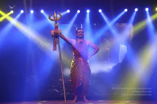 A man stands barefoot on a stage under blue and yellow spotlights, holding a tall golden trident (trishul) upright in his right hand while his left hand rests on his hip. He wears a rudraksha mala around his neck, a sleeveless saffron-orange garment, and a draped golden-orange dhoti. Behind him a painted backdrop shows a waterfall and rocky landscape. He smiles broadly. Probable Swamiji is present in this stage performance, with long swept-back hair, a rudraksha mala and a trident held in ceremonial pose.