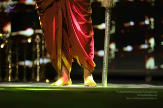 Close-up photograph of a standing person's bare feet and lower legs on a dark stage with a green-lit floor. The figure wears draped dhoti-style cloth in red and golden-yellow tones, with a gold border. Beside the feet, a tall silver staff with ornate engraved patterns and a tassel at the top stands upright. The background shows blurred stage lights, red and white bokeh, and ornamental metal fixtures.