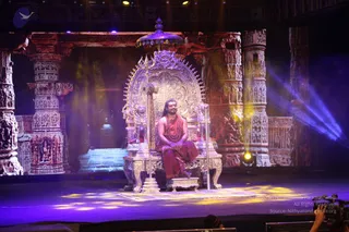 A man in orange-red robes sits on an ornate silver throne with lion-shaped armrests and a silver canopy topped by a finial, on a stage set of carved stone pillars with temple-style relief sculpture. A silver trident stands beside the throne. He wears a long rudraksha mala and a shawl over bare shoulders, and his long dark hair falls past his shoulders. Purple and blue spotlights and yellow stage lights illuminate the scene, and a camera operator is visible at the bottom edge of the frame.