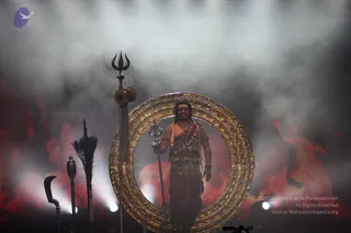 A photograph of a stage performance set in smoke with red flame projections behind. A bare-chested man stands centre within a large ornate golden circular frame, holding a trident. He wears a rudraksha-style beaded garland, a sash of beads across the chest, and rust-brown dhoti-style clothing with a wrapped waist cloth, and has a tilak on the forehead. To the left stand a trident topped staff with a drum, a curved sickle-shaped blade and a broom-like whisk on tall poles. The lighting is dramatic with spotlights low in the frame.