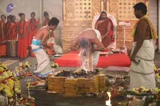 A brick-lined fire pit (homa/yagna kunda) is the centre of a ritual scene. A priest in white dhoti with a bare upper body and a purple cloth over his head bends forward over the fire, while a young assistant in white dhoti with a blue sash and garlands kneels at its edge. A shirtless man in white dhoti with a yellow-green shawl stands at right. Behind them, several women in orange-saffron sarees stand with folded hands. In the background a seated figure in orange-red robes with long hair sits on a white and red throne before a golden ornate backdrop, a red carpet below. Marigold garlands, brass vessels, fruit and flower offerings surround the fire pit.