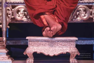 Close-up photograph of a person's bare feet resting on an ornately carved golden-brown wooden footstool with floral and scroll relief and lion-paw feet. The person wears red-orange robes with gold-bordered hems, and the upper body is cropped out of frame. Behind is a gilded carved throne-like structure with scrollwork and a dark stage backdrop. The feet are visible but the person's face and identity are not shown, so no a person candidate can be confirmed from appearance.