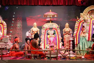 A stage puja scene before a red velvet curtain and a dark red backdrop with a glowing city-like graphic. In the centre sits a deity murti in pink and gold attire with a crown, framed by a red, white and purple floral garland arch and a gold ornamental umbrella. Gold female attendant statues flank the central figure, and a large gold figure on the right is draped in a floral garland. Three men in orange-saffron cloth with bare upper bodies and tied hair sit on the stage in front of the deity, one with orange head wrap, and perform rites with brass vessels and offerings of fruit and lamps. A brass railing separates the stage from the viewer.
